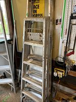 Two aluminum step ladders, one taller than the other, standing in a garage. Both have multiple wide steps and folding design.