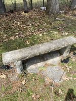 Stone bench viewed from an angled side showing stone leg and seat with rough edges on the outdoor ground with fallen leaves.