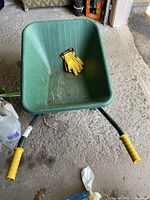 Green Tufx wheelbarrow with yellow grips on handles and yellow gloves inside, showing wear and slight rust spots.