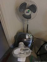 Three fans placed together: a tall Super brand pedestal fan, a square floor fan, and a tabletop fan seen in dim lighting.