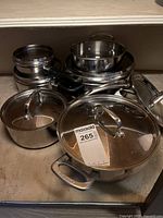 Multiple stainless steel cookware pieces including pots with lids and nested pans without lids arranged under a shelf; lids and pots have polished finish.
