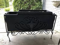 Front view of black wrought iron outdoor planter box with decorative front panel and raised legs on concrete surface