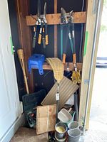 Full view of yard equipment stored in a wooden shed including hand tools, broom, rubber boots, step stool, and watering cans.