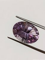 Close-up photo showing the amethyst gemstone held by tweezers, displaying the oval cut faceted surface and purple color.