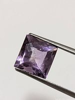 Close-up image of a purple square-cut amethyst gemstone held in tweezers, showing facets and internal clarity.