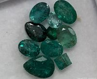 Close up photo of 10 mixed cut emerald gemstones on a white surface.