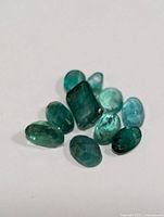 Close-up photo of nine small polished loose emerald gemstones in varying green shades on a white surface.