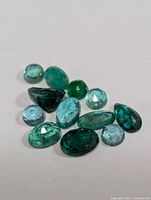 Close-up photo of 12 assorted green and bluish-green emerald gemstones, faceted and varying in cut and size laid on a white surface.