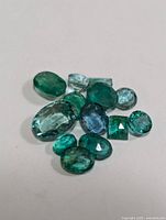 Close-up image of 11 assorted cut emerald gemstones in varying green hues and shapes.