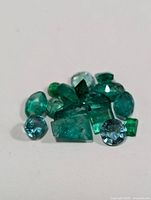 Close-up image of multiple small loose emerald and blue gemstones in various cuts and shapes on white background.