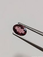 Close-up photo of an oval faceted garnet gemstone held by tweezers on a plain background.