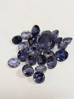 Multiple faceted iolite gemstones in various sizes and shapes shown on a neutral surface. Stones vary in violet-blue coloring and clarity.