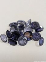 Photo of 16 loose faceted iolite gemstones varying in shape and size in shades of blue and violet on white background.