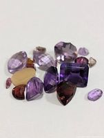 Photo showing collection of multi-colored faceted gemstones including purple amethyst variety, reddish garnet, pale yellow stone, and others in various shapes and sizes.