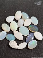 Top-down photo of 21 polished genuine opal cabochons displayed on a black surface showcasing color and shapes.