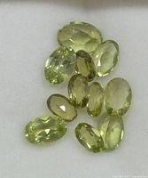 Twelve oval cut light green peridot gemstones arranged on a white background for appraisal showing their faceted cuts and clarity.