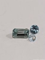 Three blue zircon gemstones displayed on a white background, showing different cuts and colors (blue-green emerald cut, light blue round brilliant, nearly colorless round brilliant).