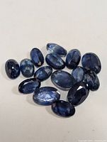 Multiple genuine loose sapphire gemstones arranged on a white surface showing faceted oval and round shapes with deep blue coloration and some internal color zoning and inclusions.