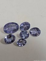 Six loose genuine tanzanite gemstones arranged on a white surface, showing various sizes and oval faceting under natural light.