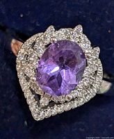Close-up front view of the silver ring showing the purple oval amethyst center stone surrounded by two rows of clear cubic zirconia stones in a scalloped design on a dark blue background.