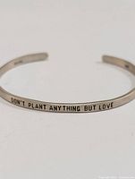 Front view of silver open cuff bangle bracelet showing the engraved phrase 'DON'T PLANT ANYTHING BUT LOVE'.