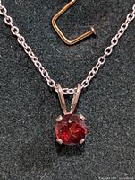 Close-up front view of the silver chain necklace with round faceted deep red garnet pendant secured by four prongs on a black background with part of a clasp visible.