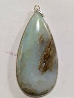 Front view of teardrop-shaped pendant showing light green, white, and brown natural gemstone with smooth polish and silver bezel setting with loop.