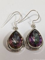 Front view of the pair of silver earrings with pear-shaped mystic topaz stones carved with leaf design and braided silver trim.
