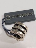 Close-up photo of silver multi-band circular pendant with Currency brand tag attached.
