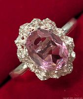 Close-up of silver ring with oval pink faceted gemstone, and finely detailed silver floral halo setting on red background.