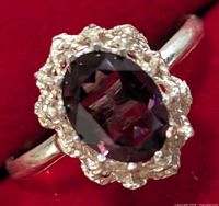 Close-up image of a silver ring featuring an oval purple faceted gemstone set in a textured silver bezel, shown on a red background.