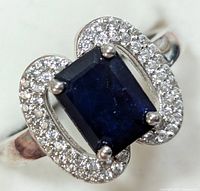 Close-up of ring front showing rectangular dark blue sapphire center stone with surrounding cubic zirconia accents on silver band.