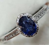 Close-up front angle of silver ring with oval blue sapphire center stone and surrounding small clear stones on band.