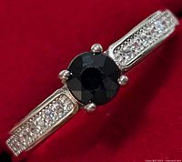 Close-up image of silver ring's top view showing black sapphire center and rows of small clear cubic zirconia stones on the band.