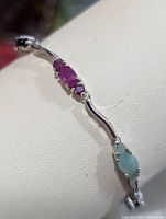 Close-up view of silver bracelet showing prong-set oval ruby and emerald gemstones on curved silver links.