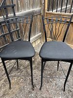 Two black metal bar stools with decorative leaf motif backs and padded black fabric seats, positioned outdoors on stone surface.