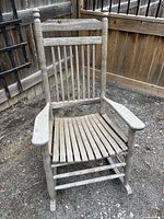 Front view of weathered solid wood rocking chair showing spindle back, armrests, and slatted seat.