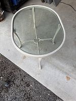 Top and side view of oval steel cream-coloured table with clear glass top on concrete ground.