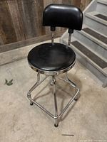 Black swivel drafting stool with round padded seat and backrest, chrome metal base and footrest, shown on a concrete floor against wood panel wall and staircase.
