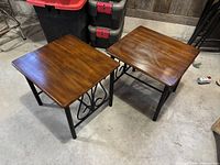 Photo showing both end tables with wooden tops and black metal legs, placed side by side on a concrete floor, wood tops display slight wear and tear.