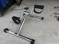 Photo showing a chrome metal sitting bicycle exercise machine with black foot pedals and straps, adjustable resistance knob on top, and rubberized feet for stability.