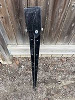 Top-down view of black metal fence post spike holder against soil and wooden fence background.