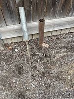 Two metal firework launching tubes secured by adjustable metal stakes set into soil along a wooden fence.