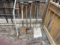 Five antique garden tools with wooden handles and rusted metal parts leaning against wooden fence on dirt ground.