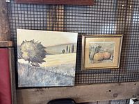 Photo showing two framed pictures hanging on a rustic wooden wall. Left is a larger square landscape painting, right is a smaller framed image of gourds.