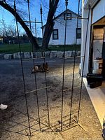 Two black wrought iron freestanding trellis standing upright outdoors on gravel with a dog in the background.