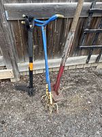 Three gardening tools lined up against a wooden fence showing full length of tamper, weed puller and dirt loosener