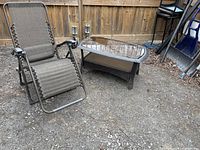 Outdoor gravity chair with gray fabric and metal frame, armrests, reclined position on gravel surface near wood fence.