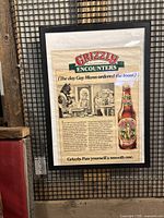 Frontal view of the framed Grizzly Beer advertisement poster hanging against a mesh/panel background.