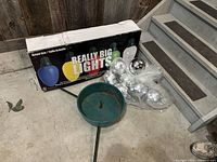 Photo showing the industrial green Christmas tree stand, boxed oversized multicolor Christmas lights labeled 'Really Big Lights,' and silver ball ornaments in plastic packaging on a concrete floor near stairs.